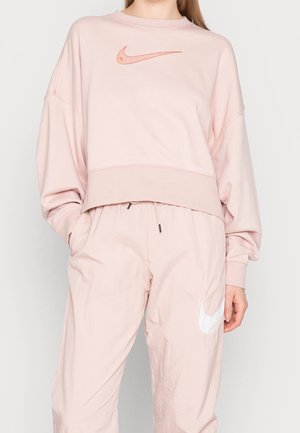 Vrouw draagt lichtroze Nike sweatshirt en bijpassende trainingsbroek met geborduurde en gedrukte Nike swoosh-logo's.