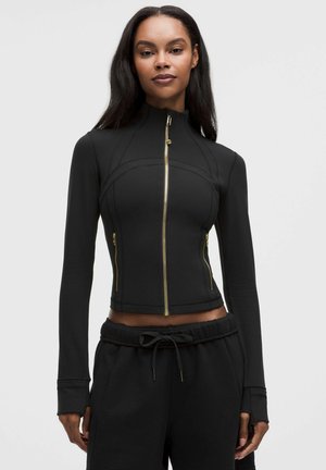Vrouw draagt een aansluitend zwart jasje met gouden ritsen en zwarte broek met trekkoord, staand tegen een effen lichte achtergrond.