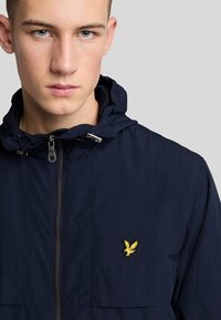 Marineblå zip-up jakke med hætte, lavet i et tekstureret stof, med en metallisk lynlås og et gult broderet logo på venstre bryst.