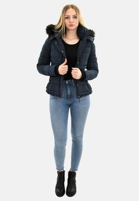 Joven mujer de pie, vistiendo una chaqueta acolchada azul marino con capucha de piel, camiseta negra, jeans ajustados azul claro y botas de tobillo negras.