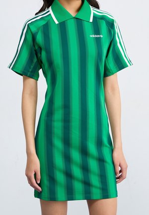Vrouw draagt een groene korte mouw Adidas polo jurk met verticale strepen en witte schouderstrepen, staand tegen een effen achtergrond.