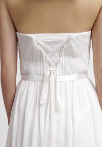 Robe blanche plissée avec un design bustier sans bretelles ; comprend une fermeture à lacets au dos et une ceinture en satin soulignant la taille.