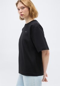 Νεαρή γυναίκα με κοντά ξανθά μαλλιά, που φοράει μαύρο T-shirt BOSS και ανοιχτόχρωμο μπλε τζιν, στέκεται πλευρικά μπροστά σε απλό φόντο.