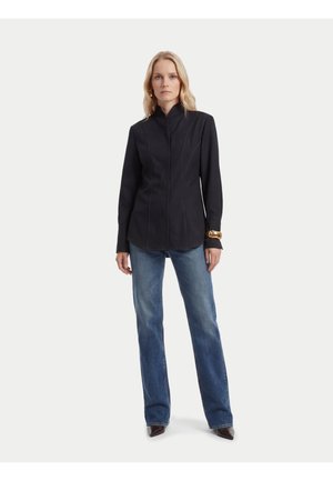 Femme blonde debout, portant une veste noire ajustée, un jean bleu, des chaussures marron pointues et un bracelet en or à son poignet gauche.