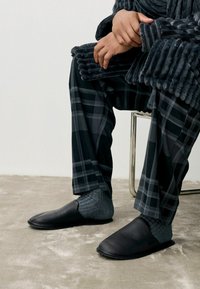 Black leather slip-on slippers with a smooth texture, worn with grey ribbed socks, complemented by plaid-patterned pants in dark tones.