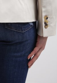 Main reposant à côté de la poche d'un jean en denim bleu foncé, portant une manche de blazer beige avec trois boutons visibles.