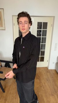 Mladý muž s tmavými kučeravými vlasmi zapína čiernu pruhovanú košeľu, stojí v miestnosti s drevenou podlahou, bielymi stenami a sklenenými dverami.