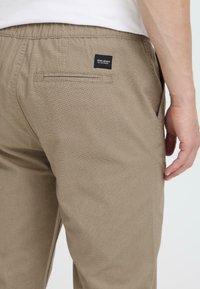 Pantalones caqui en una tela texturizada con cintura elástica. Presentan un bolsillo trasero con una pequeña etiqueta negra y un detalle de costura mínima.