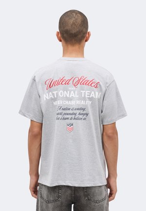 Homme aux cheveux courts portant un t-shirt gris avec l'inscription « United States Men's National Team » et un logo bouclier des États-Unis dans le dos.