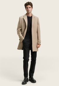 Abrigo de lana beige con un diseño entallado, frente abierto, cuello sobredimensionado y puños abotonados. Combinado con pantalones negros y botas negras gruesas.
