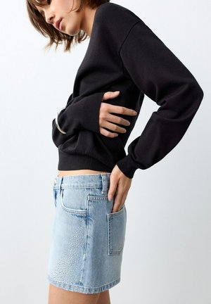 Donna che indossa una maglia nera a maniche lunghe e una minigonna di jeans azzurri con dettagli di strass, in piedi di lato con le mani sui fianchi.