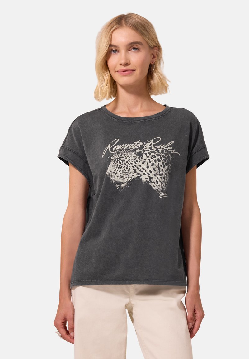 Femme portant un T-shirt noir délavé avec un motif tête de léopard et le texte "Rewrite Rules", assorti à un pantalon beige clair.
