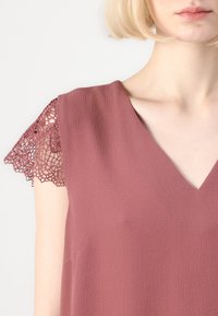 Bluzica ružičaste boje s V-izrezom i čipkastim detaljima na kratkim rukavima, izrađena od teksturiranog materijala. Glatko drapiranje, elegantan dizajn.
