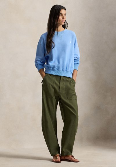 Ανοιχτό μπλε βαμβακερό φούτερ με στρογγυλή λαιμόκοψη και ριμπ μανσέτες, σε συνδυασμό με χαλαρής εφαρμογής παντελόνι σκούρου πράσινου και καφέ σανδάλια. Μινιμαλιστικός σχεδιασμός.