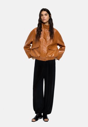Kobieta stoi na tle jednolitego tła, ubrana w brązową skórzaną kurtkę bombowca z przednimi kieszeniami, luźne czarne spodnie i czarne sandały z odkrytymi palcami.