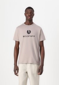 Light beige cotton t-shirt with short sleeves, featuring a black "BELSTAFF" logo and an emblem above it. Smooth texture and relaxed fit.