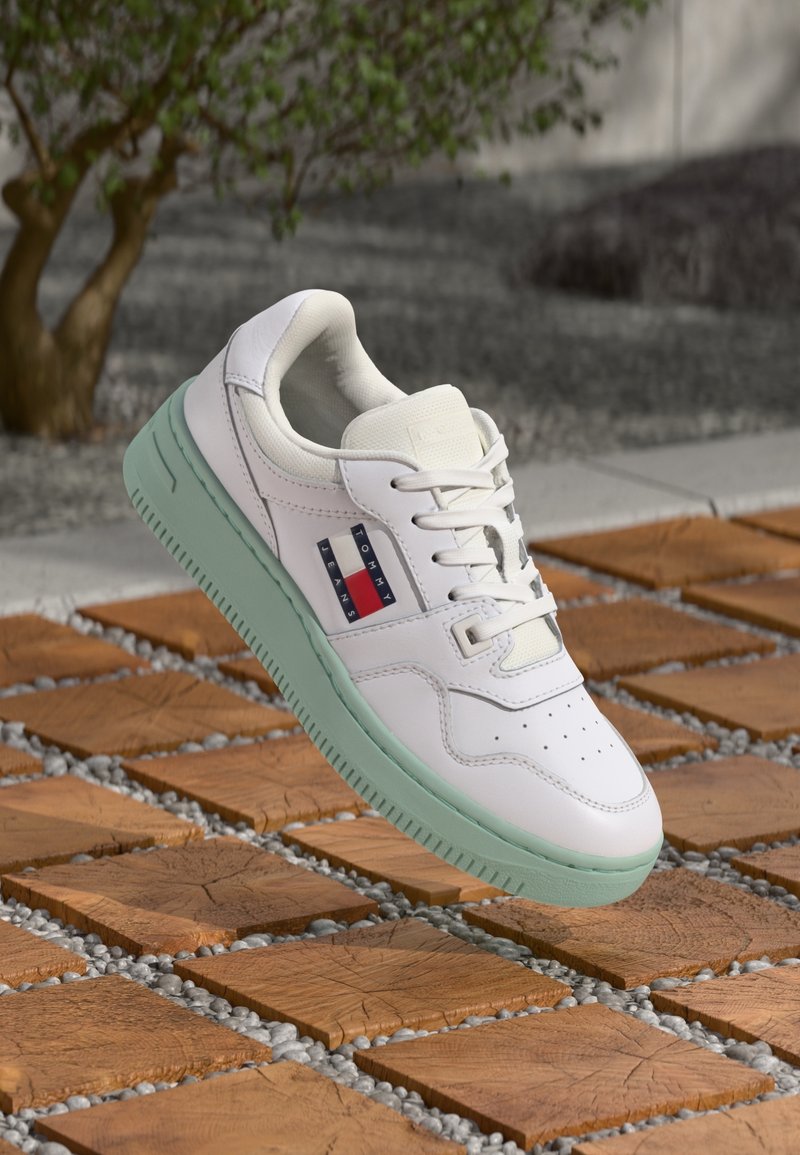 Basket blanche avec semelle vert clair, logo Tommy Jeans sur le côté, lacets noués, présentée sur un carrelage en bois et un sol de cailloux en extérieur.