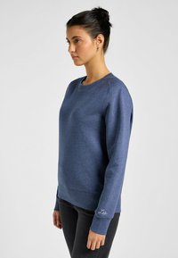 Granatowy sweter z okrągłym dekoltem, długimi rękawami i ściągaczami; ma subtelnie haftowane logo na lewym rękawie.