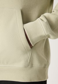 Main dans la poche avant d'un sweat-shirt beige clair avec poignets et ourlet côtelés, montrant la texture du tissu et les détails des coutures.
