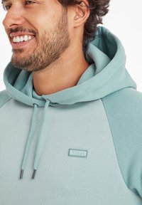 Close-up of a smiling man wearing a light teal hoodie with a visible "TOG24" logo on the chest.