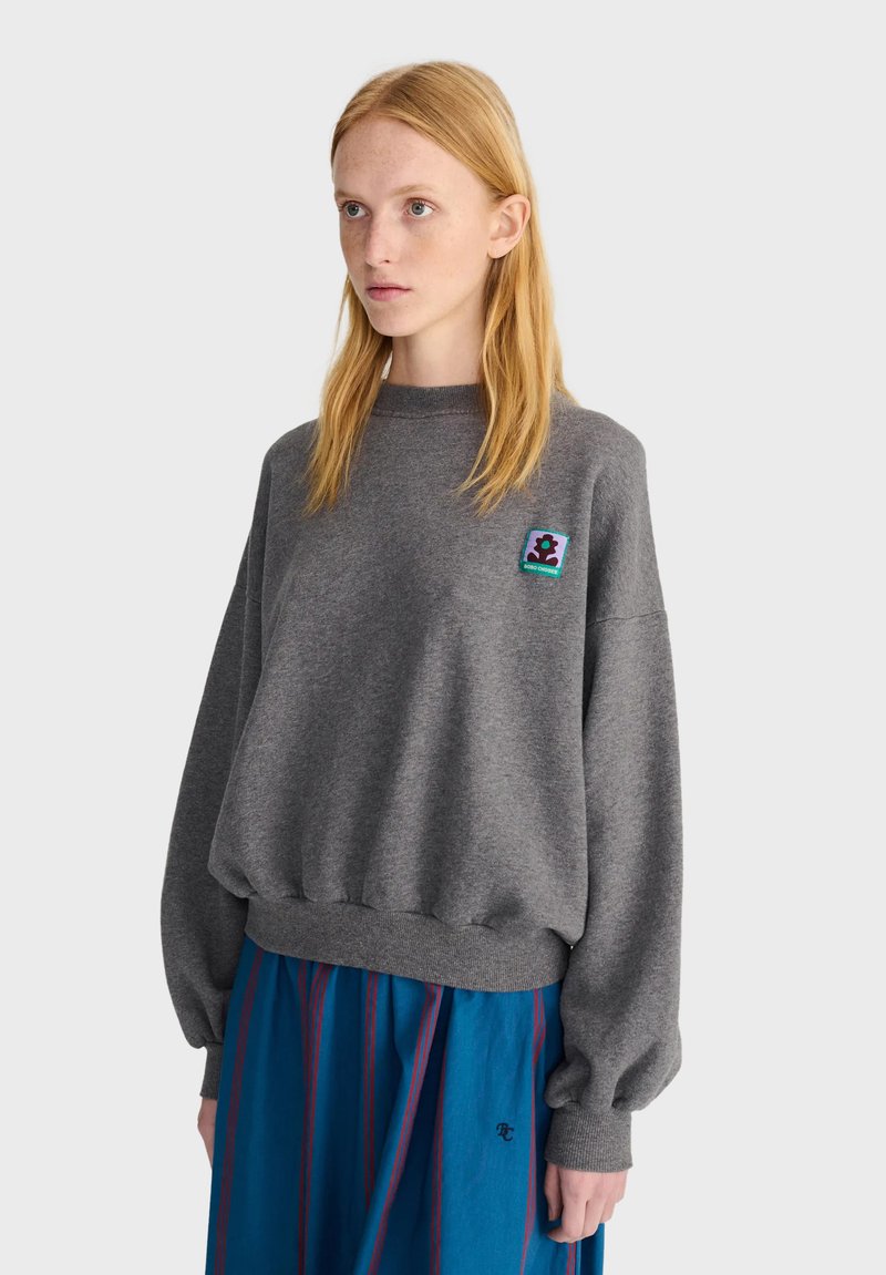 Jeune femme aux cheveux blonds raides portant un sweat-shirt gris ample avec un petit patch coloré et une jupe bleue à rayures rouges.