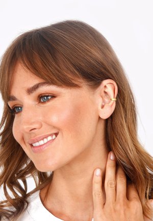 Guldørering i hoop-stil på en models øre, med et glat, cirkelformet design. Modellen har brunt hår, lys hud og neutral makeup, og smiler.