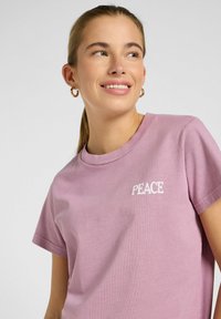Camiseta de algodón de color rosa claro, con mangas cortas, cuello redondo y la palabra "PAZ" impresa en blanco en el pecho. Textura suave y corte relajado.