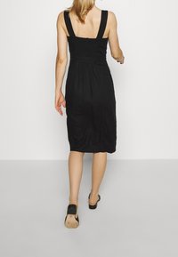 Svart klänning med breda axelband, smockad liv, längd till mitten av vaden, flödande design och texturerad tyg. Bärs med svarta slip-on skor.