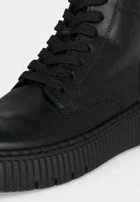 Μαύρο δερμάτινο sneaker ψηλού τύπου με παχιά ραβδωτή λαστιχένια σόλα και μαύρα υφασμάτινα κορδόνια, εμφανίζεται σε κοντινό πλάνο σε λευκό φόντο.