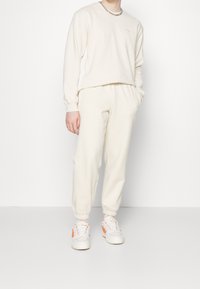 Sudadera color crema y joggers a juego con puños acanalados, combinados con zapatillas blancas con acentos naranjas y un collar de cadena.