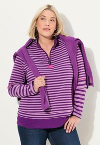 Lila gestreiftes Oberteil mit einem Reißverschlusskragen, kombiniert mit einem einfarbigen lilafarbigen Pullover, der über die Schulter gelegt ist. Lässiger Stil mit weichem Stoffgefühl.