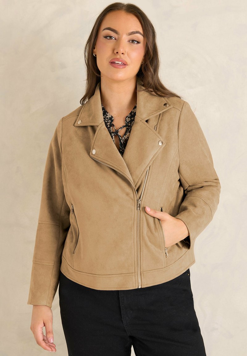 Femme aux longs cheveux bruns portant une veste motard en daim beige par-dessus un haut noir à fleurs et un pantalon noir, debout devant un fond neutre.