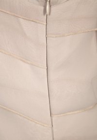 Indumento in tessuto beige con un design testurizzato e a strati. Presenta delle cuciture delicate lungo le cuciture e una chiusura con zip sul retro.