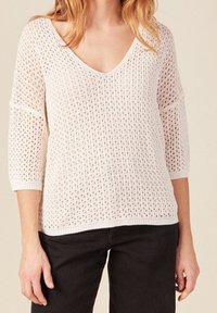 Bílý pletený svetr s volnou, otevřenou tkaninou, výstřihem do V a rukávy o délce 3/4. Dokončený žebrovanými okraji. Spárováno s černými kalhotami.
