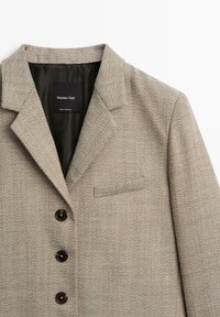 Béžový tvídový blazer s výřezovými klopami, čtyřmi černými knoflíky, náprsní kapsou a hladkou podšívkou. Načal složený uvnitř.