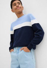 Teen che indossa un maglione a blocchi di colore blu, bianco e navy, con le mani nelle tasche dei jeans azzurri, che guarda leggermente in alto e sorride.