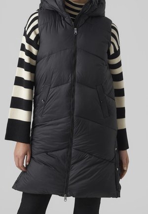 Veste longue sans manches noire matelassée avec fermeture éclair frontale et fentes latérales à bouton-pression, portée sur un pull rayé noir et blanc.