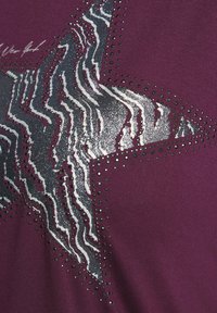 Kiiltävä hopea ja musta tähti, jossa on aaltoilevia raidoja ja pieniä mustia pisteitä purppuraisella kankaan taustalla.