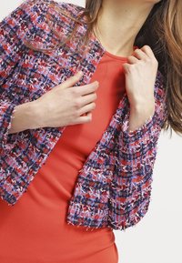 Flerfärgad vävd jacka i röd, blå och vit, med fransiga kanter. Bärs över en enfärgad orange topp med en strukturerad yta.