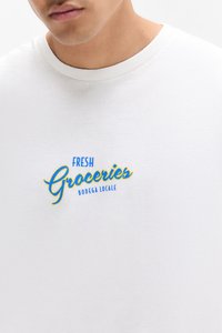 White t-shirt with a round neck, featuring blue and yellow embroidered text that reads "FRESH Groceries Bodega Locale."