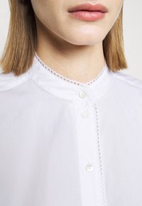 Chemise blanche boutonnée avec un col montant festonné et une délicate bordure en dentelle le long de la patte de boutonnage, réalisée en tissu lisse avec un léger éclat.