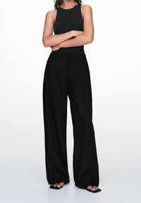 Débardeur noir moulant associé à un pantalon large noir. Tissu lisse, taille haute et longueur cheville. Sandales glissantes texturées.