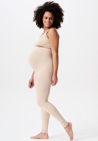 Beige graviditetstopp och leggings-set, mjukt elastiskt tyg, figurnära design, slät textur, bärs av en person med lockigt hår.