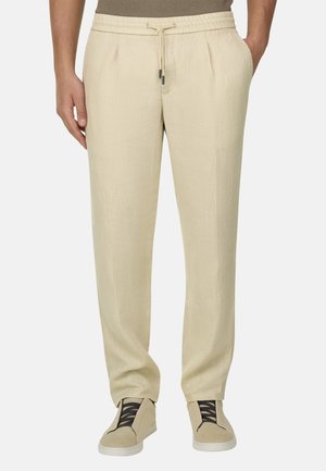 Pantalones beige con cordón usados con zapatillas a juego de color beige con cordones negros, manos descansando casualmente en los bolsillos.