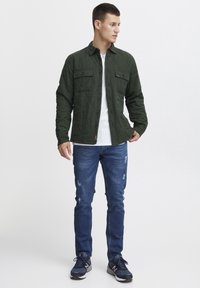 Quiltet olivengrønn skjortjakke med to brystlommer og knappelukking, kombinert med slitte blå jeans og marineblå sneakers.