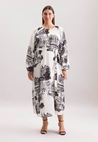 Langes weißes Kleid mit schwarzem architektonischem Muster, langen Ärmeln, betonter Taille und Kordelzug am Ausschnitt. Kombiniert mit schwarzen Riemchensandalen.