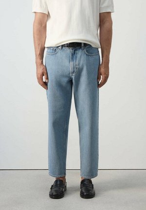 Hombre con vaqueros de pierna recta azul claro, cinturón negro, camisa blanca de manga corta y mocasines negros, de pie sobre suelo gris.