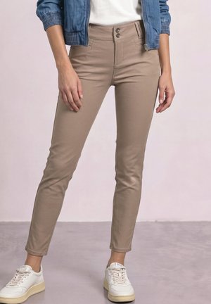 Personne portant un pantalon beige slim, des baskets blanches, un haut blanc et une veste en jean bleue sur un fond uni.