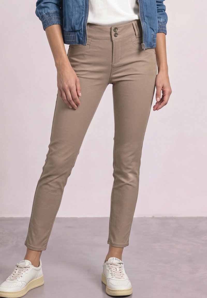 Personne portant un pantalon beige slim, des baskets blanches, un haut blanc et une veste en jean bleue sur un fond uni.