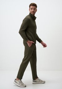 Ensemble de sport vert olive comprenant un haut zippé avec un col haut et un pantalon fuselé assorti. Le matériau semble lisse, avec un design ajusté.